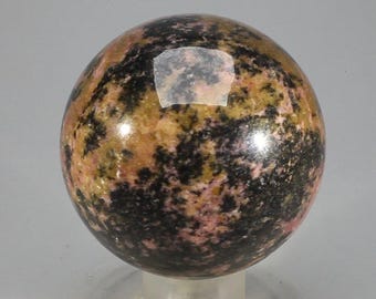 May include: A polished sphere with a mottled pattern of black, pink, and tan. The gemstone ball is displayed on a clear, plastic stand. The sphere's surface has a glossy finish, reflecting light.