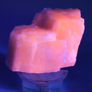 May include: A raw, crystalline mineral specimen exhibiting a vibrant, glowing orange hue under ultraviolet light. The stone features a jagged, multi-faceted surface with sharp edges and a dense, opaque texture, resting upon a small, clear circular stand.