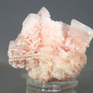May include: A close-up of a pink mineral specimen. The mineral has a layered, crystalline structure with a light pink hue. The base of the mineral is a lighter, more granular texture. The specimen is mounted on a clear stand against a grey background.