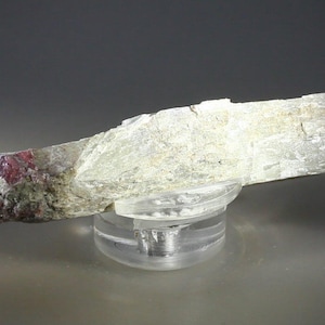 Op de afbeelding: Een langwerpig mineraal specimen in lichte kleuren, mogelijk een kristal, rust op een heldere, cirkelvormige displaystandaard. Het mineraal heeft een ruw, getextureerd oppervlak met gebieden in lichtroze en wit. De standaard is gemaakt van helder plastic.