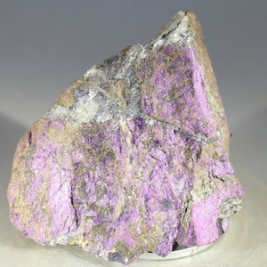 May include: A rough, gray and purple rock with a crystalline texture. The rock has a purple hue with a metallic sheen.