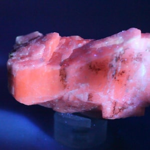 May include: A raw, crystalline mineral specimen displaying a vibrant, fluorescent coral-pink hue under ultraviolet light. The structure features angular, blocky formations with visible dark, mineralized inclusions embedded within the surface texture.