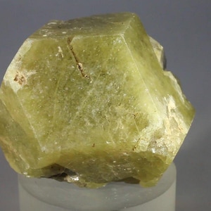 May include: A raw, pale yellow-green mineral specimen with a geometric, multifaceted crystalline structure. The surface displays natural fractures, internal inclusions, and varying levels of translucency across its angular, blocky form.