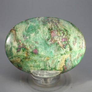 Puede incluir: Una piedra preciosa pulida de forma ovalada que presenta una mezcla vibrante de patrones fibrosos de color verde claro y oscuro, intercalados con inclusiones distintas e irregulares de color magenta en toda la superficie, exhibida sobre un soporte de plástico transparente.
