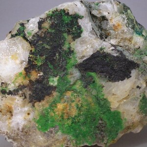 Puede incluir: Primer plano de una roca con depósitos minerales verdes y negros. La roca tiene una textura blanca y cristalina.