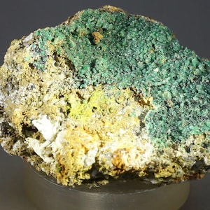 Puede incluir: Primer plano de un espécimen mineral con una superficie rugosa y texturizada. El mineral presenta una vibrante capa verde cristalina sobre una base de tonos amarillos, beiges y blancos. La muestra se exhibe sobre un fondo neutro.