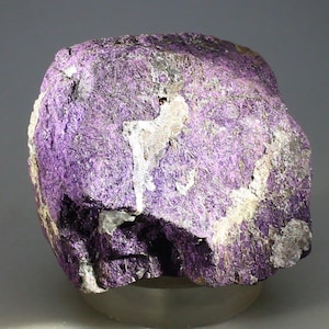 May include: A rough-hewn, natural stone specimen with a vibrant purple hue. The rock's surface displays a mix of textures, with areas of crystalline structure and patches of lighter, almost white, mineral deposits. The overall shape is irregular, showcasing its raw, unpolished state.