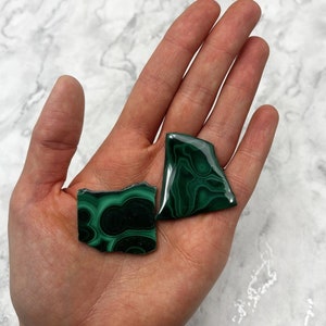 May include: Two green malachite stones with swirling patterns. The stones are irregular in shape and have a smooth, polished surface.