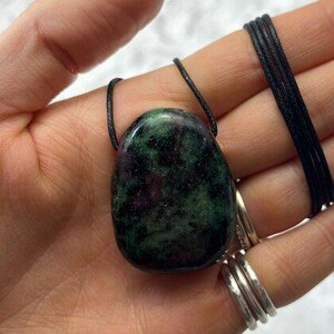 May include: An oval gemstone pendant necklace with a mix of green and maroon colours. The stone is suspended from a black cord. The necklace is held in a hand.
