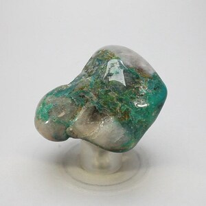 May include: A polished, natural stone with a rounded shape. The stone features a mix of colors, including shades of green, teal, and gray. The stone is mounted on a clear, circular stand, showcasing its unique texture and color variations.