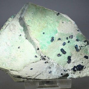 May include: A rough-hewn mineral specimen with a light green and white surface, speckled with small, dark blue-black crystalline formations. The rock has a textured appearance, with variations in color and surface detail.