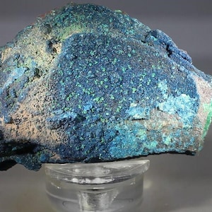 May include: A raw mineral specimen featuring a textured surface with deep blue and vibrant turquoise crystalline clusters. The rock displays a rough, uneven formation with varying shades of blue and green, resting on a clear acrylic display stand.