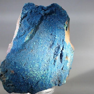 May include: A raw mineral specimen featuring a vibrant, deep blue crystalline surface with a textured, granular appearance. The rock displays natural variations in color intensity, with small patches of lighter blue and green mineral deposits.