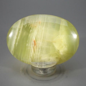 May include: An oval-shaped light green stone cabinet knob with a polished, translucent finish. The stone features natural white banding and subtle orange inclusions throughout its surface, mounted on a clear circular base with a metal screw attachment.