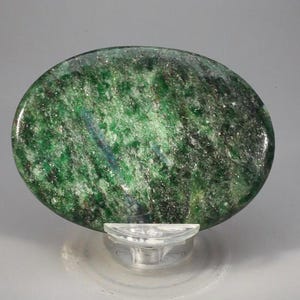 May include: A polished, oval-shaped stone with a mottled green surface, speckled with darker green and black inclusions. The stone is displayed on a clear, acrylic stand, showcasing its natural textures and colors. The overall appearance suggests a decorative or collectible item.