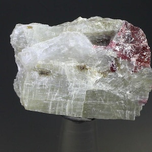 Op de afbeelding: Een doorschijnend, kristallijn mineraal specimen met een gelaagde structuur. De steen heeft een bleke, gebroken witte basiskleur met gebieden van dieprode en bruine insluitsels. Het mineraal is gemonteerd op een heldere, cilindrische standaard.