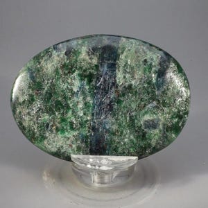 May include: A polished, oval-shaped gemstone with a mottled green and blue pattern. The stone is mounted on a clear, pedestal-style display stand, showcasing its natural textures and colors. The gemstone's surface has a mix of dark and light green hues.