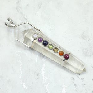 May include: A clear quartz crystal pendant with seven colorful gemstone accents. The gemstones are arranged in a row and are various colors, including purple, blue, green, orange, and red. The pendant has a silver-colored metal loop.