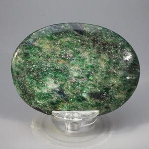May include: A round, polished stone with a vibrant green hue, speckled with darker green and black inclusions. The stone is mounted on a clear, plastic display stand, showcasing its natural beauty. The surface has a glossy finish, reflecting light.