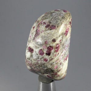 May include: A polished, rounded stone featuring a mottled white and grey base with embedded, irregular deep pink and magenta crystal inclusions throughout its surface. The stone has a smooth, glossy texture and a curved, asymmetrical shape.