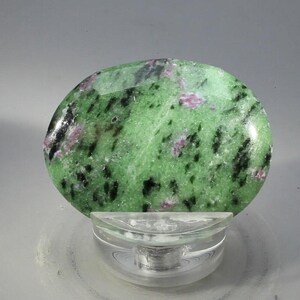 May include: A polished, oval gemstone with a vivid green hue, speckled with black and pink markings. The stone is presented on a clear, circular display stand. This mineral specimen is a striking example of natural beauty, perfect for collectors.