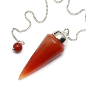 Può includere: Un pendolo di cristallo di corniola rossa con una catena d'argento e una piccola perla rossa. Il pendolo ha la forma di un cono e ha una superficie liscia e lucida.