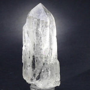 Puede incluir: Un cristal de cuarzo transparente con una parte superior puntiaguda y una superficie rugosa y texturizada. El cristal es transparente, permitiendo el paso de la luz, y está sobre un fondo oscuro. Esta gema natural es un artículo decorativo.