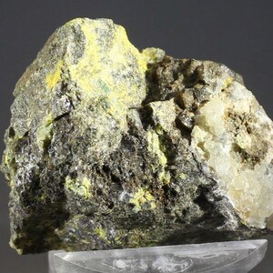 Puede incluir: Primer plano de un espécimen mineral que presenta una mezcla de colores y texturas. La roca tiene una base gris oscuro con parches de amarillo brillante y formaciones cristalinas blanquecinas. La superficie es rugosa e irregular, mostrando características geológicas naturales.