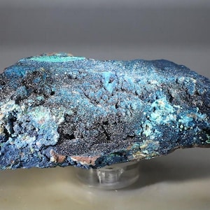 May include: A raw mineral specimen featuring a rough, crystalline texture with deep blue and vibrant turquoise coloration. The surface displays a sparkling, granular appearance with natural crevices and uneven edges, showcasing a mix of dark navy and bright cyan mineral deposits throughout the stone.