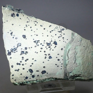 May include: A close-up of a mineral specimen. The specimen has a light gray base color with numerous dark blue, irregularly shaped inclusions scattered across its surface. The edges of the specimen have a greenish hue.