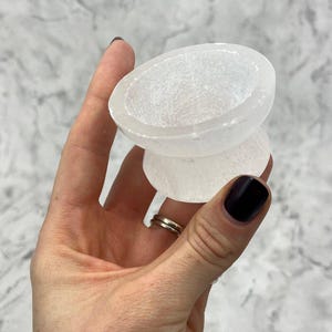 May include: A white selenite crystal bowl with a smooth, polished surface. The bowl is shaped like a bell and has a wide, shallow opening.