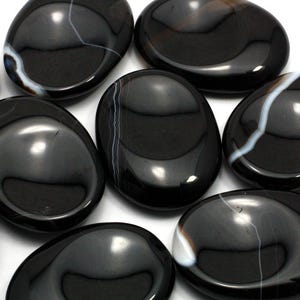 Puede incluir: Una colección de piedras de palma de ágata negra con bandas blancas. Las piedras son lisas y pulidas, y están dispuestas en un patrón aleatorio.