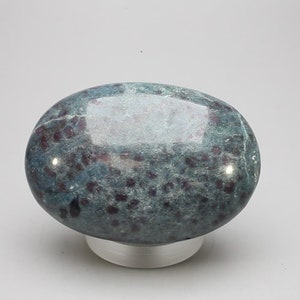 May include: A smooth, oval-shaped green and purple gemstone with a speckled pattern. The gemstone is sitting on a white stand.