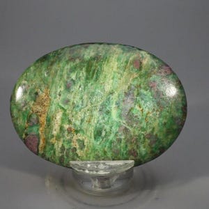 May include: An oval-shaped polished gemstone featuring a mottled surface with vibrant shades of green, interspersed with deep magenta and dark brown inclusions. The stone has a smooth, glossy finish and is displayed on a clear plastic stand.