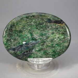 May include: A polished, oval-shaped stone with a vibrant green hue, speckled with lighter shades and dark veins. The stone is displayed on a clear, pedestal-style stand, showcasing its natural beauty. The stone's surface has a textured appearance.