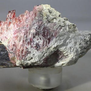 May include: A raw mineral specimen featuring a jagged, crystalline structure with deep red and translucent pink hues integrated into a rough, textured white and grey matrix. The stone displays sharp, vertical striations and uneven edges.