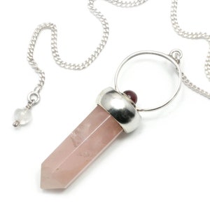 Può includere: Un pendolo di cristallo rosa con una catena d'argento e un anello d'argento in cima. Il cristallo ha una forma a punta e ha una piccola pietra rossa in cima.