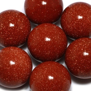 May include: Close-up of seven polished, round, reddish-brown gemstones. The stones have a speckled, glittery appearance, with tiny reflective particles throughout. The gemstones are arranged in a circular pattern against a white background.