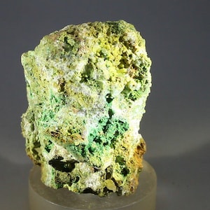 Puede incluir: Primer plano de un espécimen mineral con una superficie rugosa y texturizada. La roca muestra una mezcla de colores, incluyendo amarillo vibrante, verde brillante y toques de marrón. El mineral está montado sobre un soporte cilíndrico transparente.
