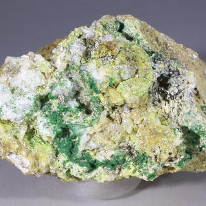 Puede incluir: Primer plano de una muestra de roca con depósitos minerales verdes, amarillos y blancos. La roca tiene una superficie rugosa y desigual.