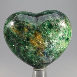 May include: A heart-shaped stone sculpture with a polished surface. The stone is primarily green with streaks of gold and dark green. The sculpture is mounted on a silver-colored metal base.