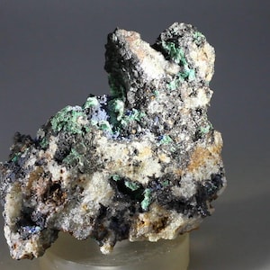 May include: A rough, gray rock with green and blue mineral deposits. The rock has a variety of textures and colors, including white, brown, and black.