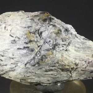 May include: A close-up of a mineral specimen with a layered, textured appearance. The rock is primarily white with streaks of black and hints of yellow and brown. The surface has a crystalline, almost fibrous quality.