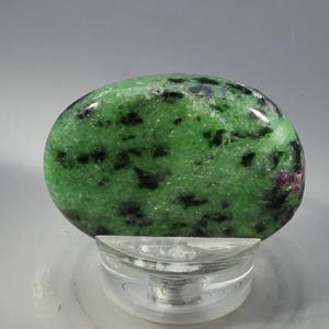 May include: A smooth, oval gemstone, mainly green with black and red markings. The stone is presented on a clear glass stand, highlighting its natural patterns. The surface of the gemstone is polished and reflects light, showing its texture.
