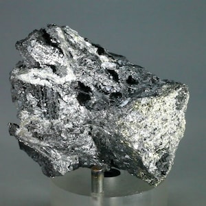 Könnte beinhalten: Ein großes, raues, graues und silbernes metallisches Mineralstück. Das Mineral hat eine kristalline Struktur und wird auf einem durchsichtigen Ständer präsentiert.