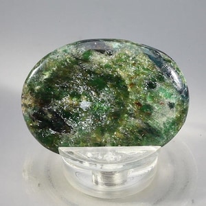 May include: A polished, oval-shaped gemstone with a vibrant green hue, speckled with lighter green and white inclusions. The stone is mounted on a clear, rounded display stand, showcasing its natural beauty. The gemstone's surface reflects light, highlighting its unique patterns.