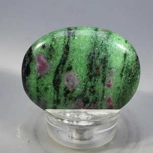 May include: A polished, oval gemstone with a green base, black veining, and red inclusions. The stone sits on a clear, circular stand. The gemstone's natural patterns are visible. A decorative item, the stone is a unique piece.