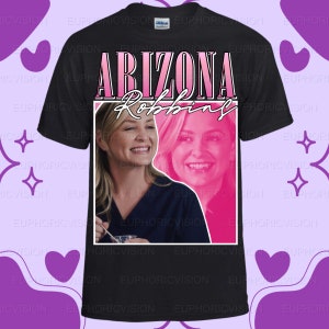 May include: Black t-shirt featuring a pink and white graphic with the text "ARIZONA Robbins" and two images of a smiling woman. The shirt is made of a soft material.