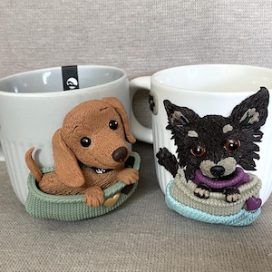Puede incluir: Dos tazas de cerámica con figuras de perro en arcilla tridimensional. La taza izquierda tiene un perro salchicha marrón en una cama verde, mientras que la derecha muestra un perro de pelo largo negro y fuego en una cama azul claro con detalles.