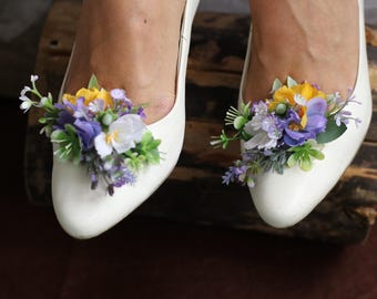 May include: A pair of white bridal shoes with a floral embellishment on each toe. The flowers are purple, yellow, and green.